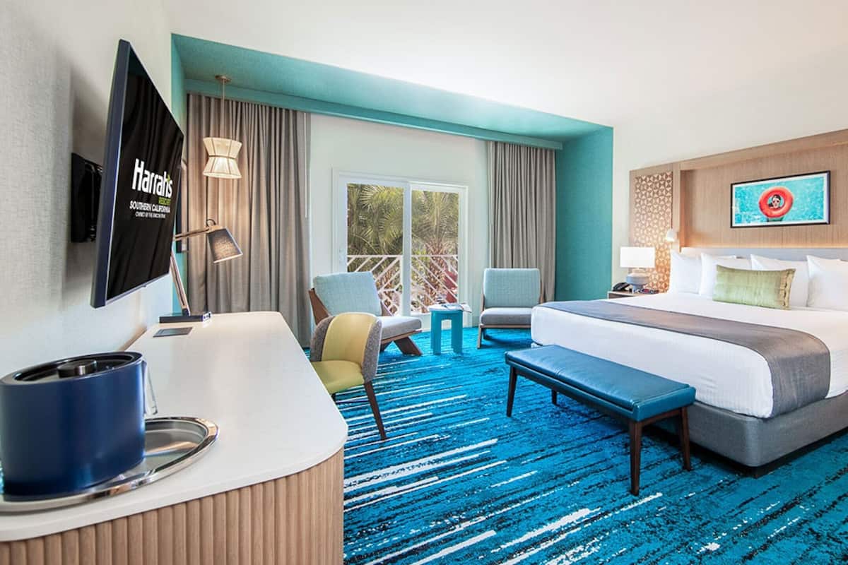 A hotel room with a large bed, a desk with a flat-screen TV, a seating area with chairs, a blue carpet, and a large window with curtains.