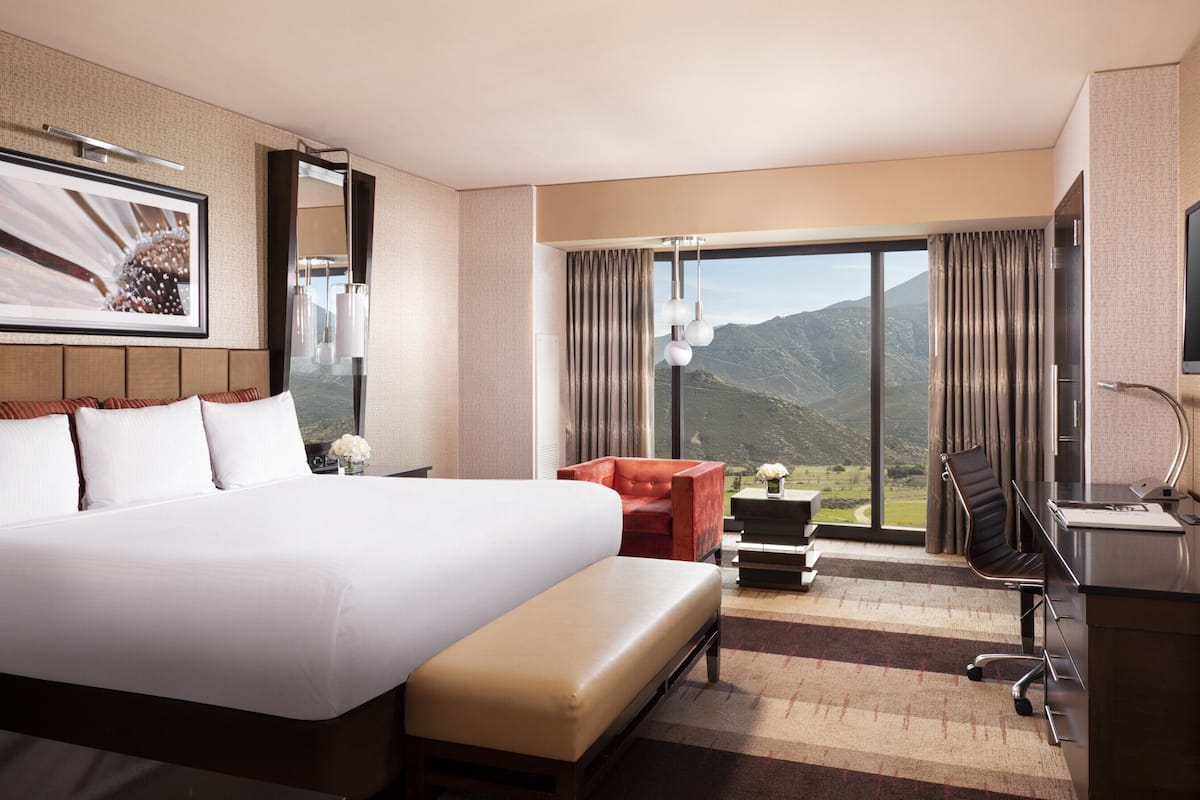 A hotel room with a large bed, a desk with a chair, a TV, and a view of mountains.