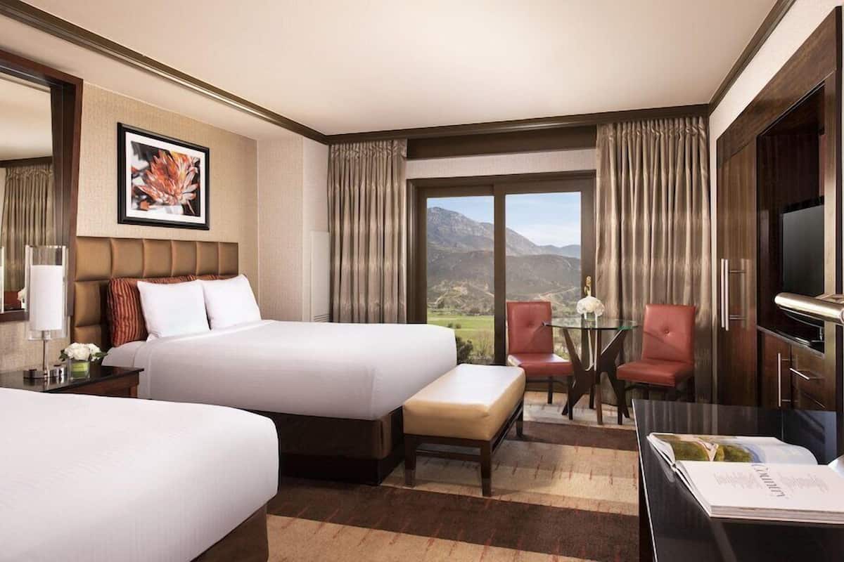 A hotel room with two beds, a desk with a chair, a TV, and a view of mountains.