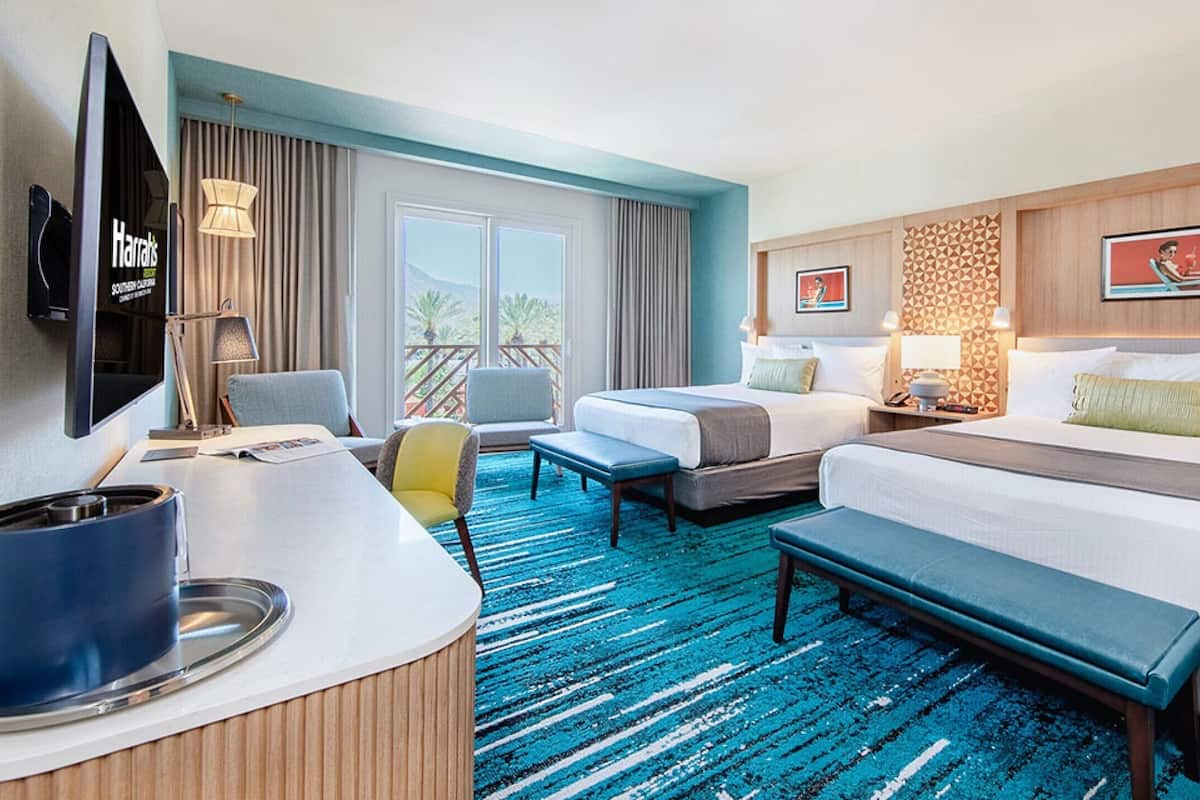 A hotel room with two beds, a desk, a chair, a TV, and a balcony with a view.