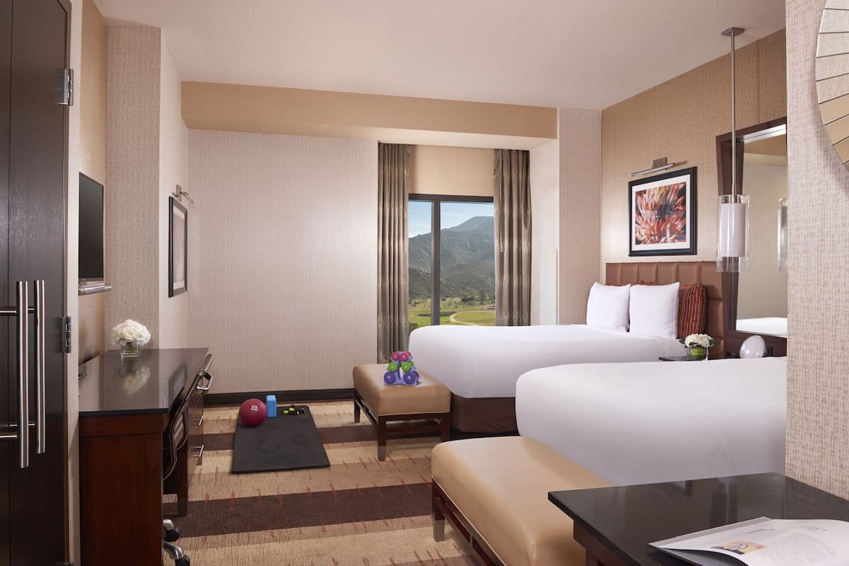 A hotel room with two beds, a desk, a chair, a TV, a window with a mountain view, and a small exercise ball.