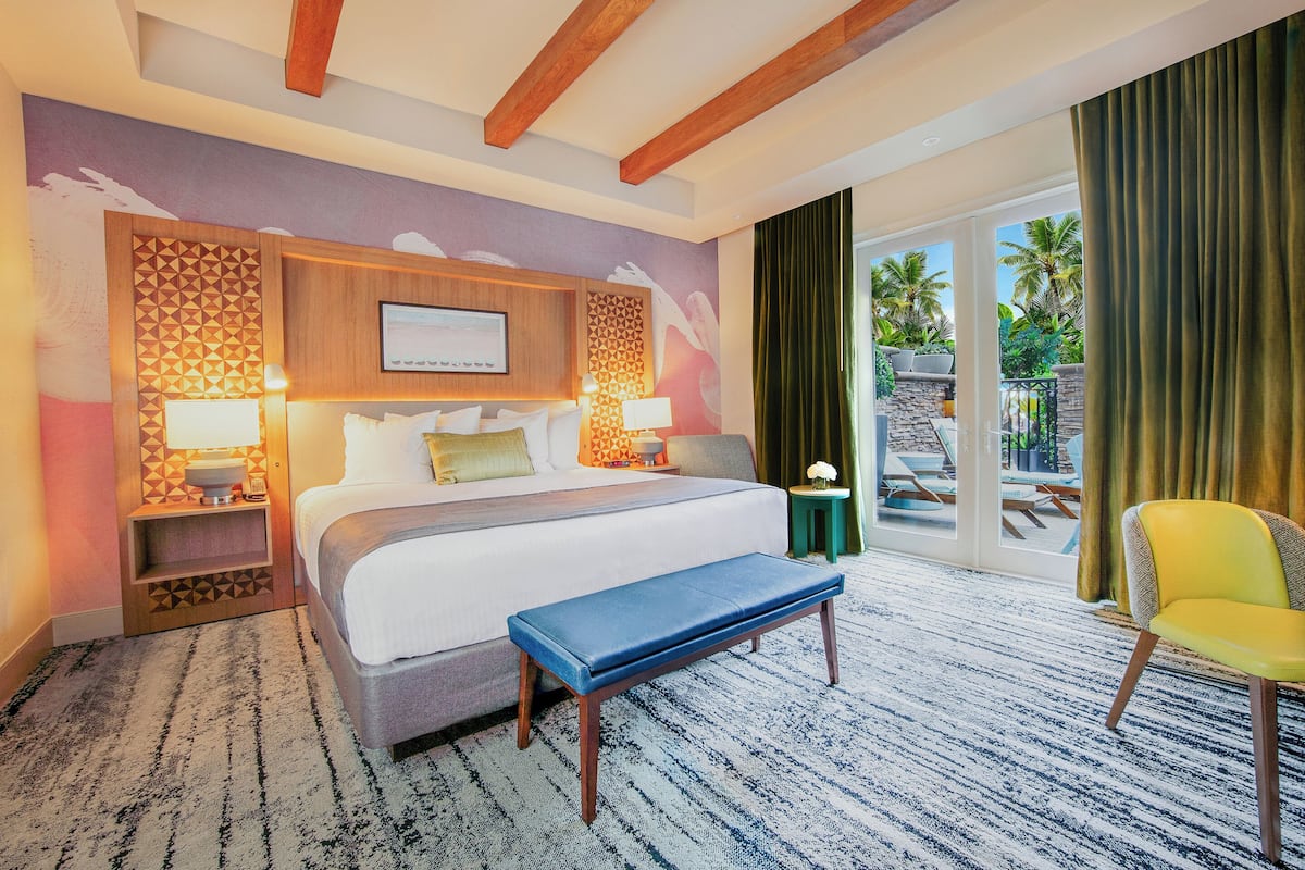 A hotel room with a large bed, a bench, a chair, and a view of palm trees through the sliding glass door.