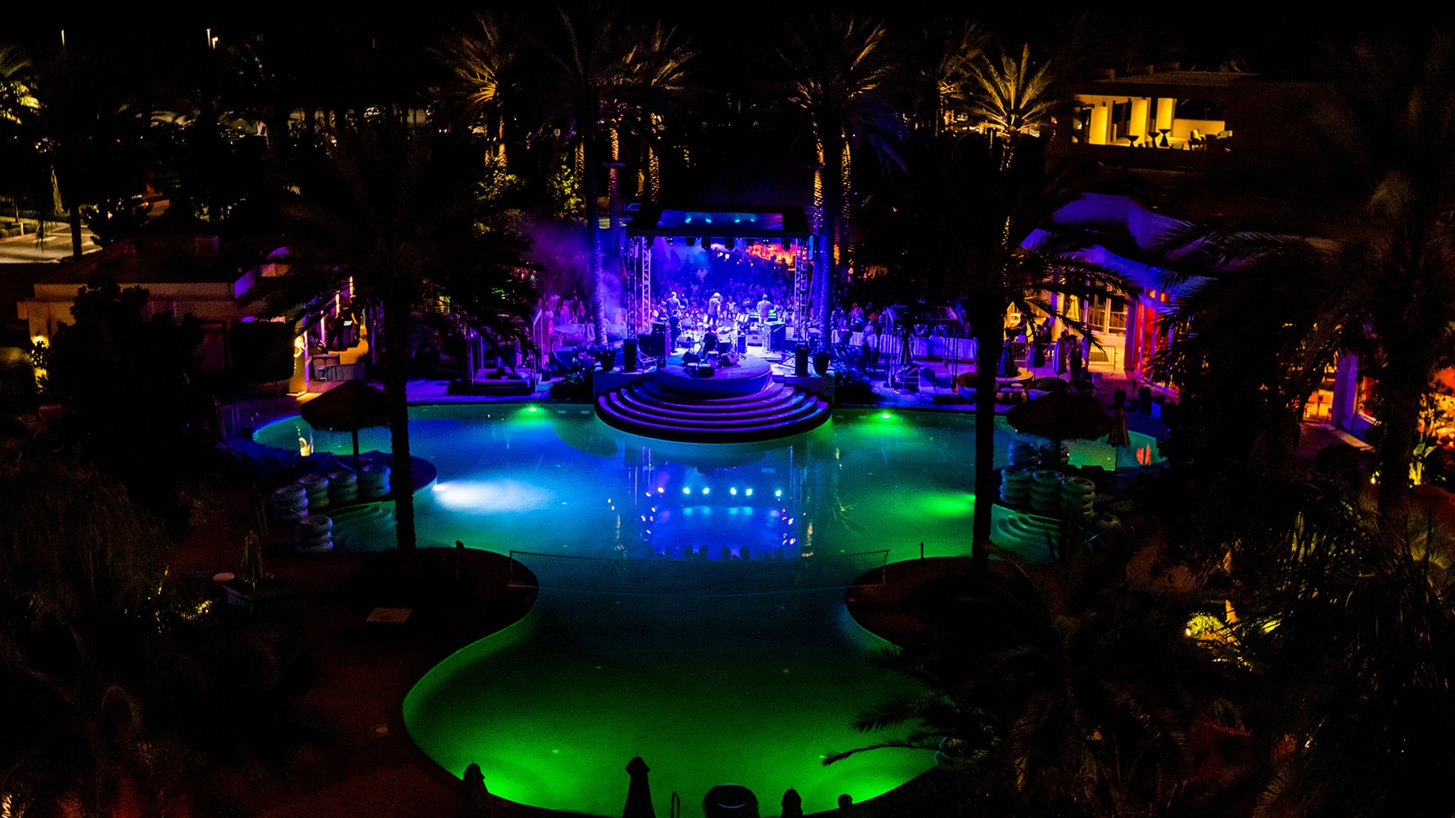A nighttime view of a resort with a central pool area, surrounded by palm trees and illuminated by colorful lights.