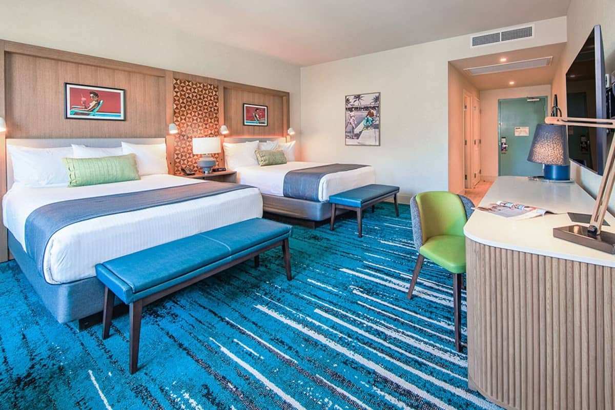 A hotel room with two beds, a bench, a desk with a chair, and a TV.