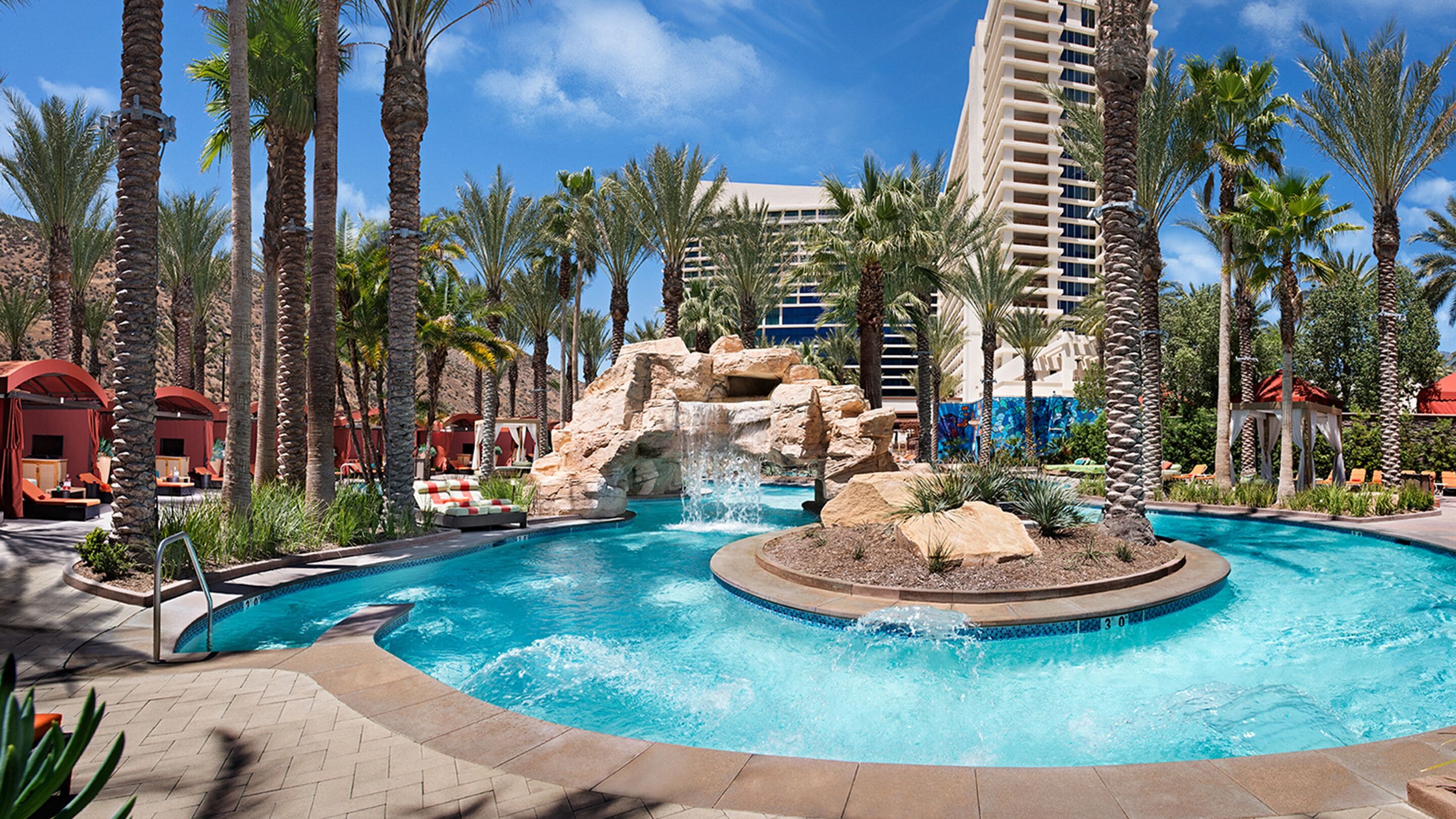 A resort pool with a waterfall, surrounded by palm trees and a multi-story building in the background.