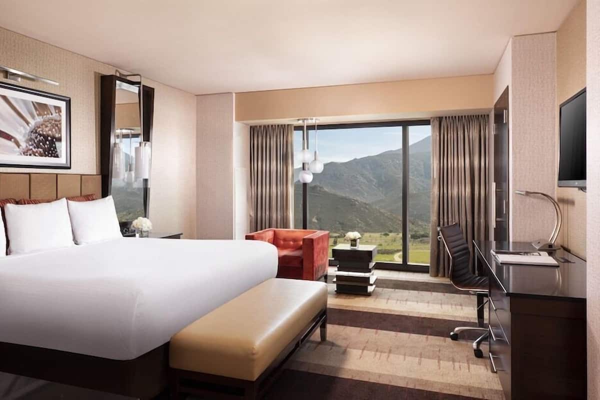 A hotel room with a large bed, a desk with a chair, a TV, and a view of mountains.