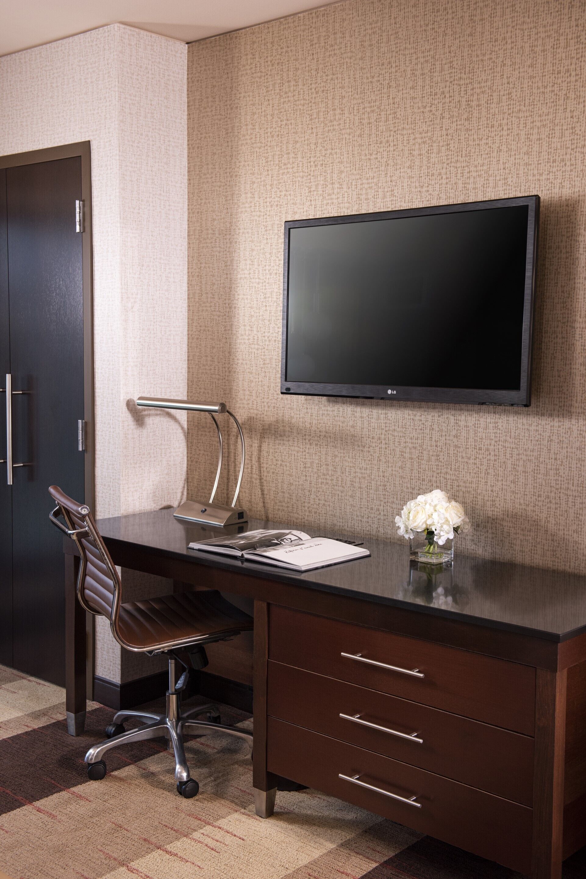 Ein Hotelzimmer mit dunklem Holztisch, einem modernen Sessel, einem Flachbildfernseher, einer kleinen Vase mit weißen Blumen und einer Tür mit Griff.