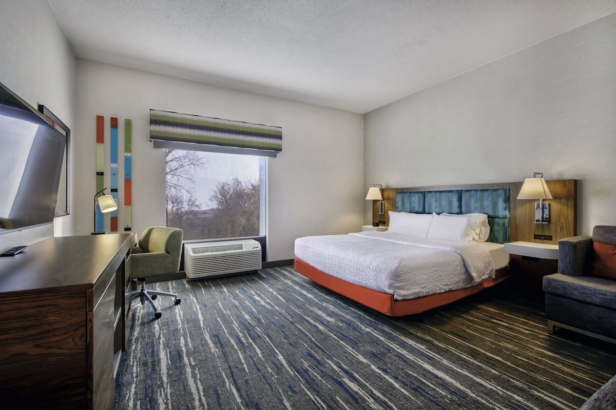 A hotel room with a bed, desk, chair, TV, air conditioning unit, and a window with a view.
