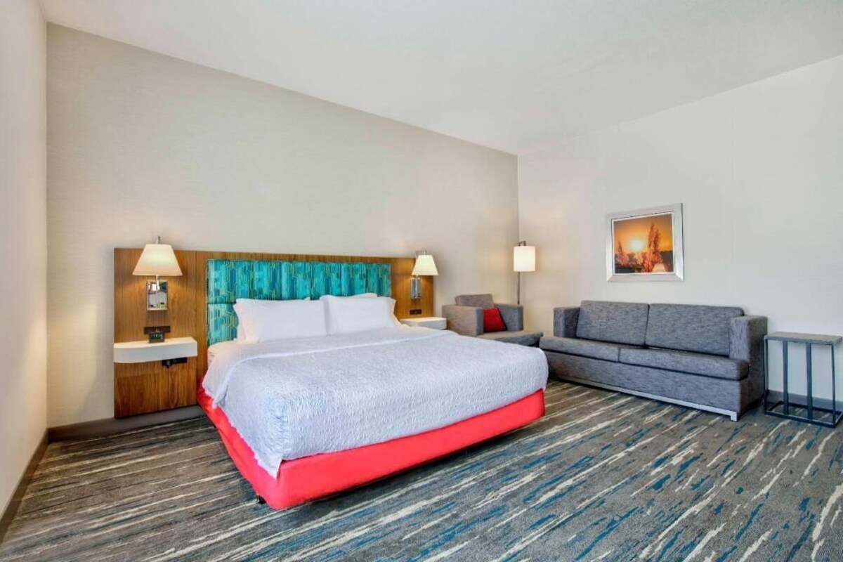 Een hotelkamer met een bed, een grijze bank, een nachtkastje en een aan de muur hangend schilderij.