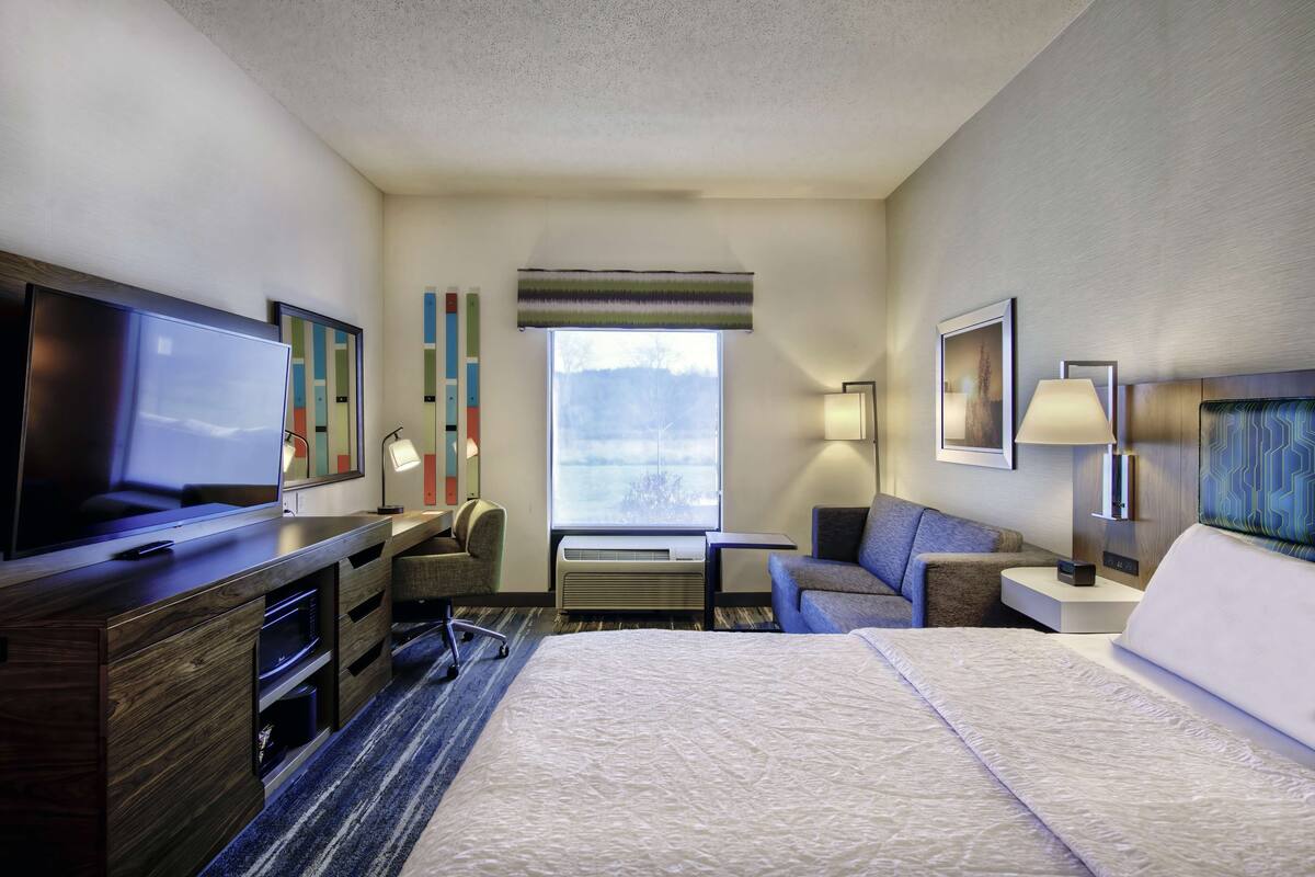 A hotel room with a bed, a desk with a flat-screen TV, a chair, a sofa, a window, and a wall-mounted lamp.