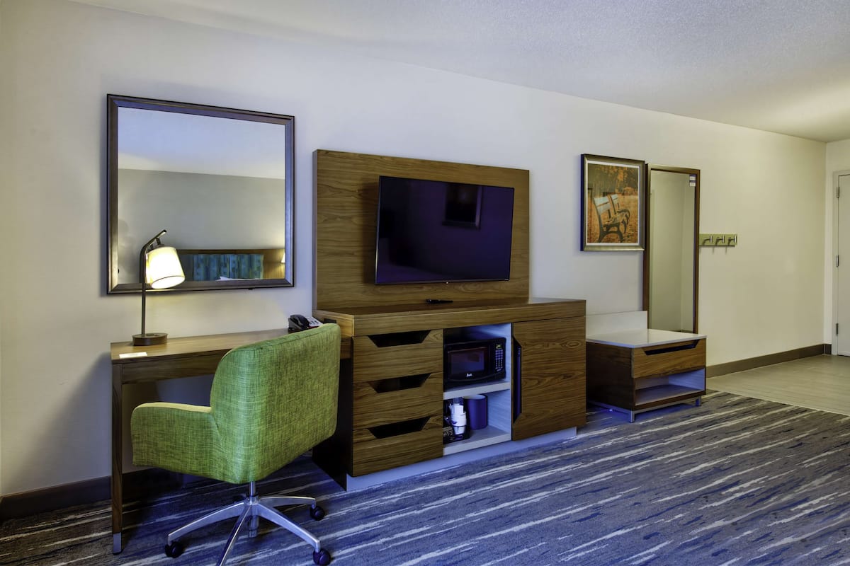A hotel room with a wooden desk, a green office chair, a flat-screen TV, a mirror, and a carpeted floor.