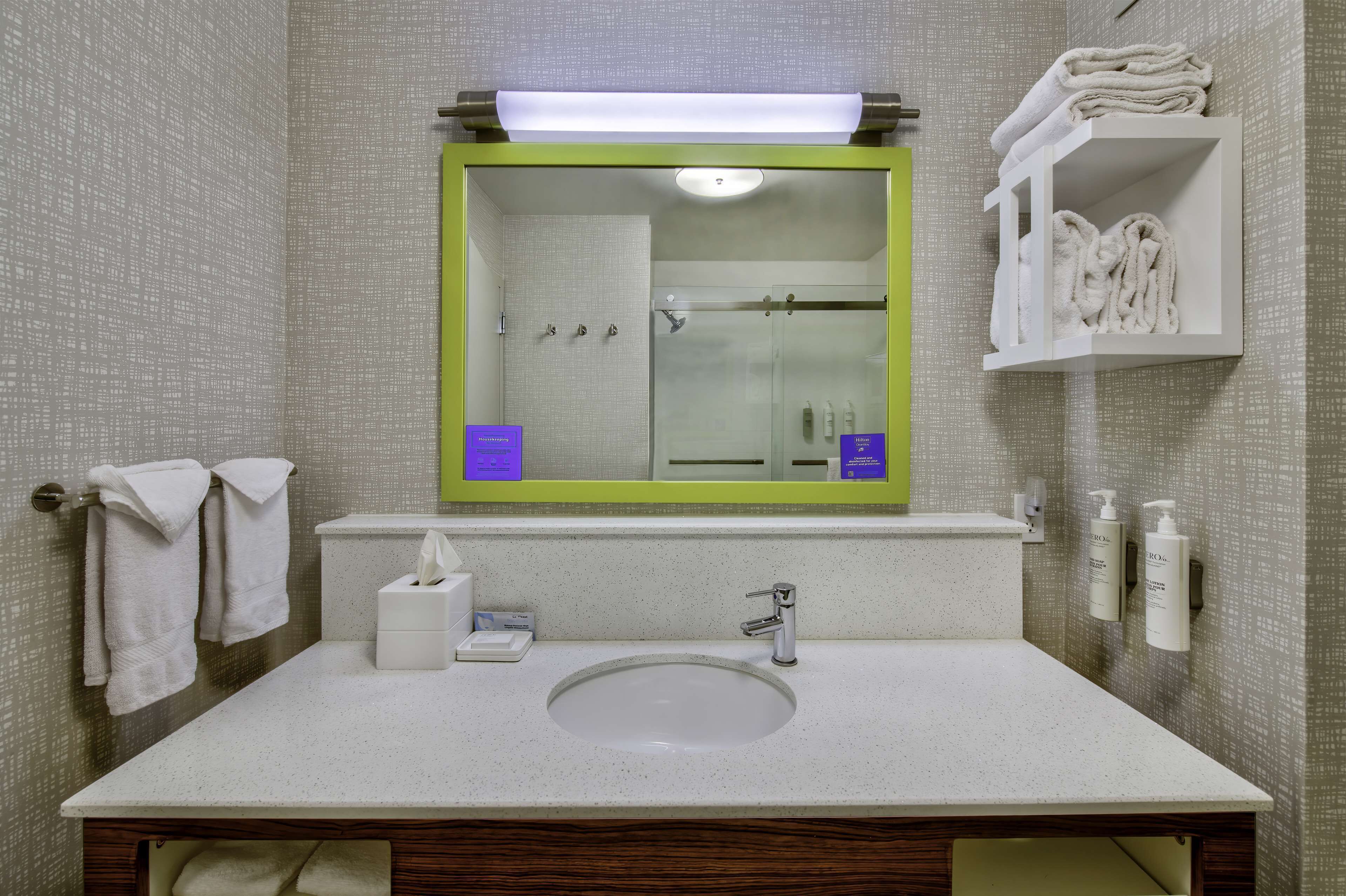A bathroom with a white sink, a mirror with a green frame, a towel rack, and a shelf with towels.