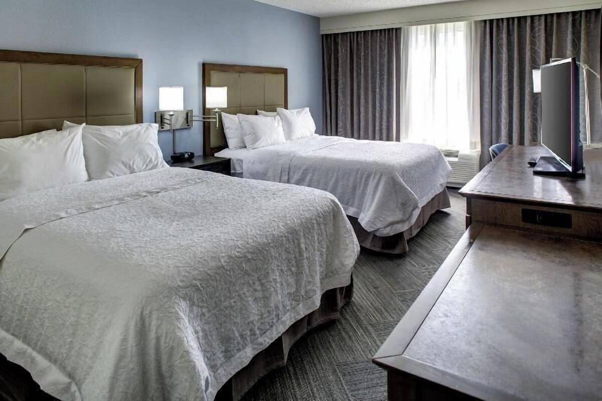 A hotel room with two beds, a desk, a television, and a window with curtains.