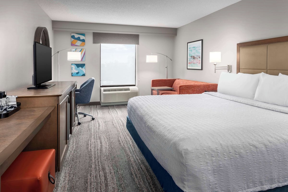 A hotel room with a large bed, a desk with a computer, a chair, a sofa, and a window with a view.