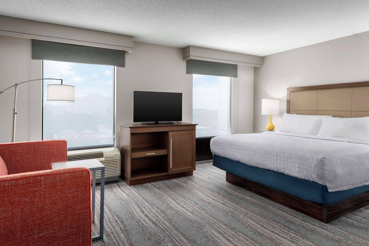 A hotel room with a bed, a TV on a stand, a chair, and a desk.