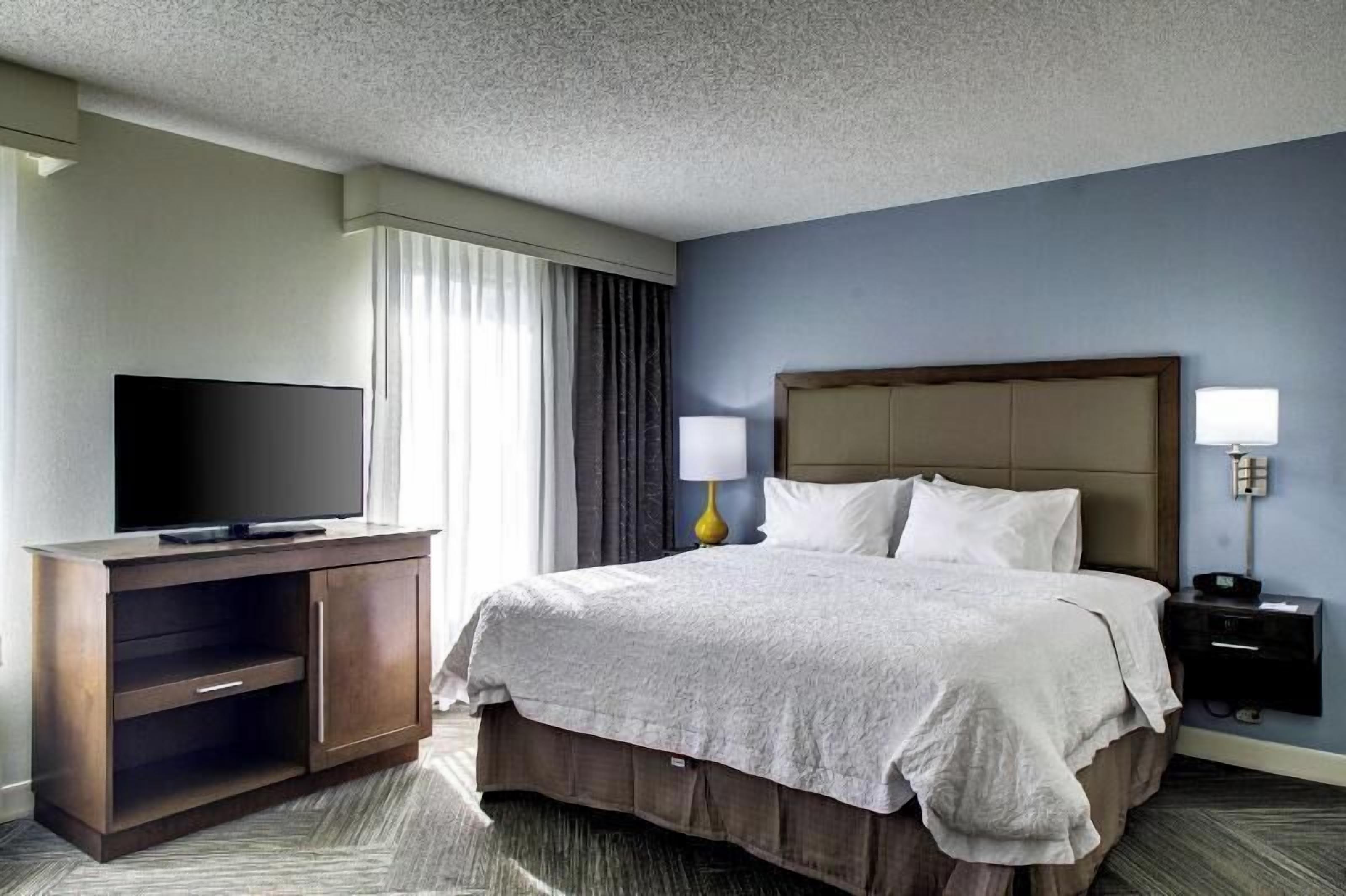 A hotel room with a bed, a TV on a wooden cabinet, a nightstand with a lamp, and a window with curtains.
