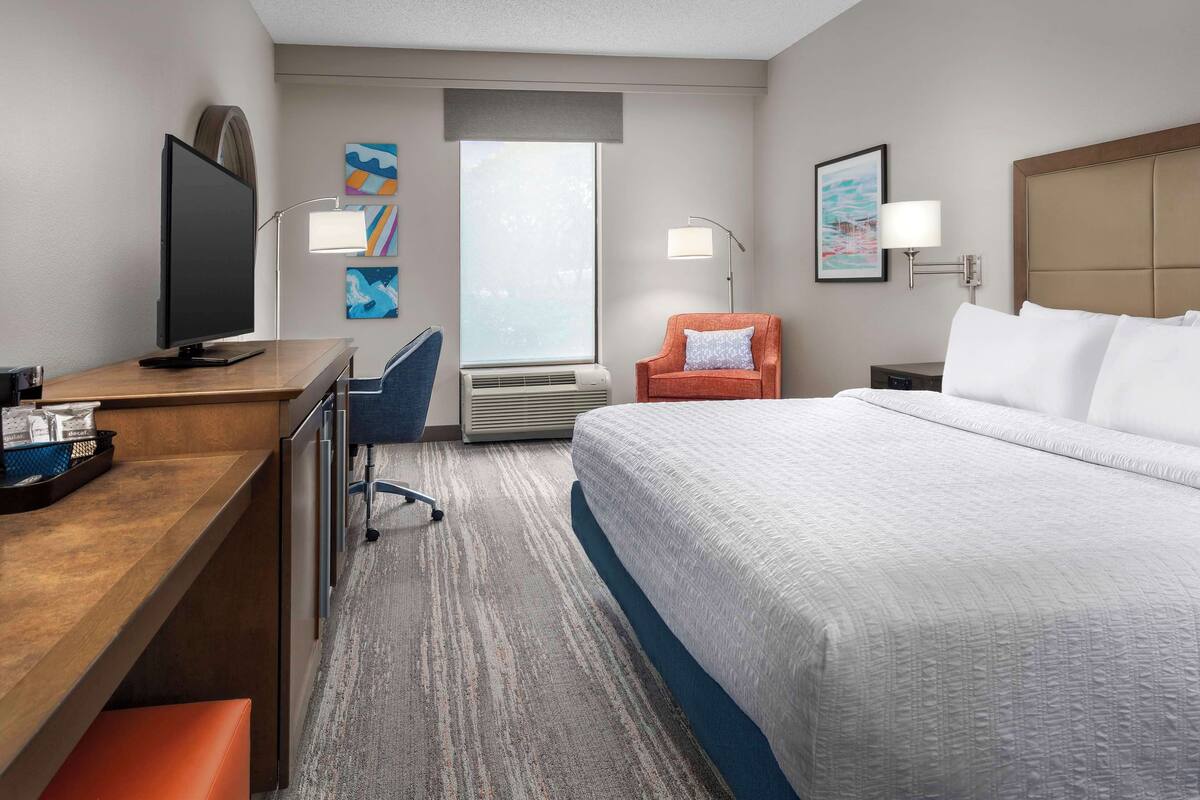 A hotel room with a large bed, a desk with a computer, a chair, a TV, and a window with a view.
