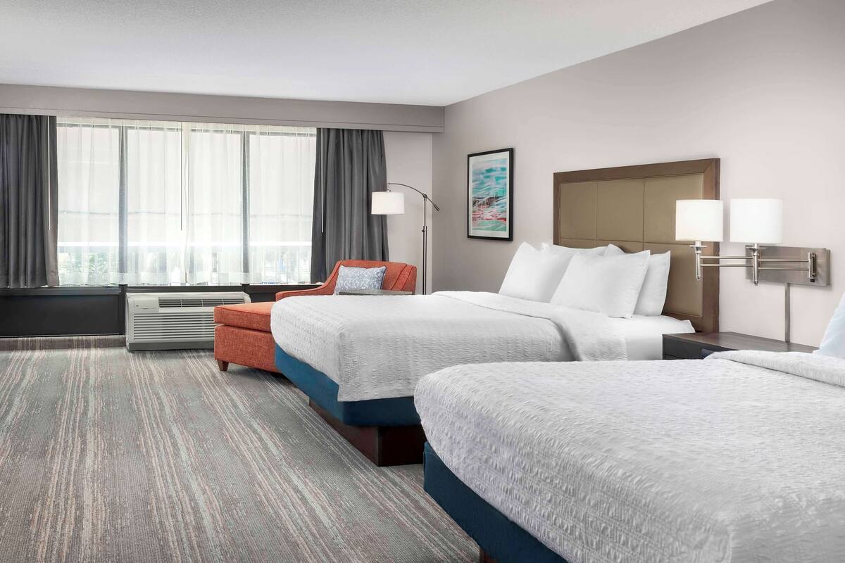 A hotel room with two beds, a sofa, a large window with curtains, a floor lamp, and a painting on the wall.