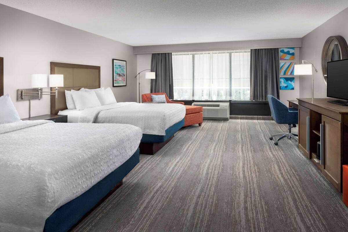 A hotel room with two beds, a desk, a chair, a TV, and a large window with curtains.