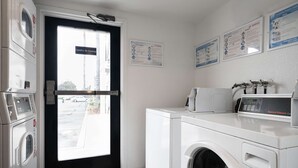 Une buanderie équipée d’un lave-linge à chargement frontal, d’un sèche-linge et d’un panneau de commande. Des panneaux explicatifs sont affichés sur le mur, ainsi qu’un panneau indiquant qu’il est interdit de fumer.
