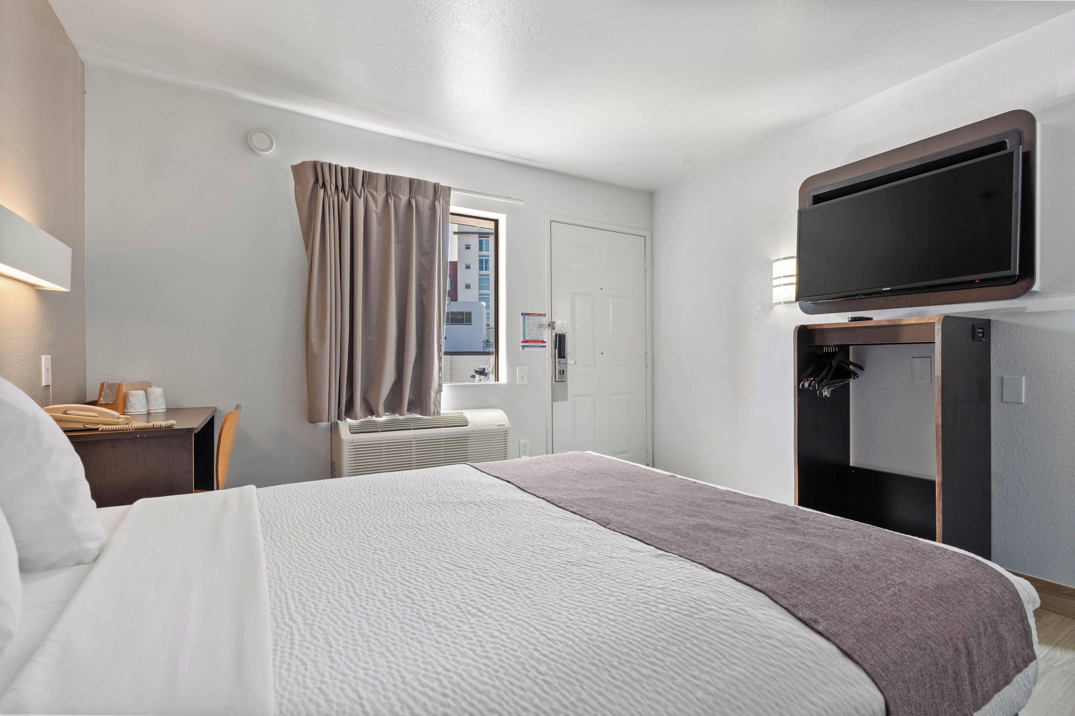 Quarto de hotel com cama, escrivaninha com telefone, televisão na parede e janela com cortinas.