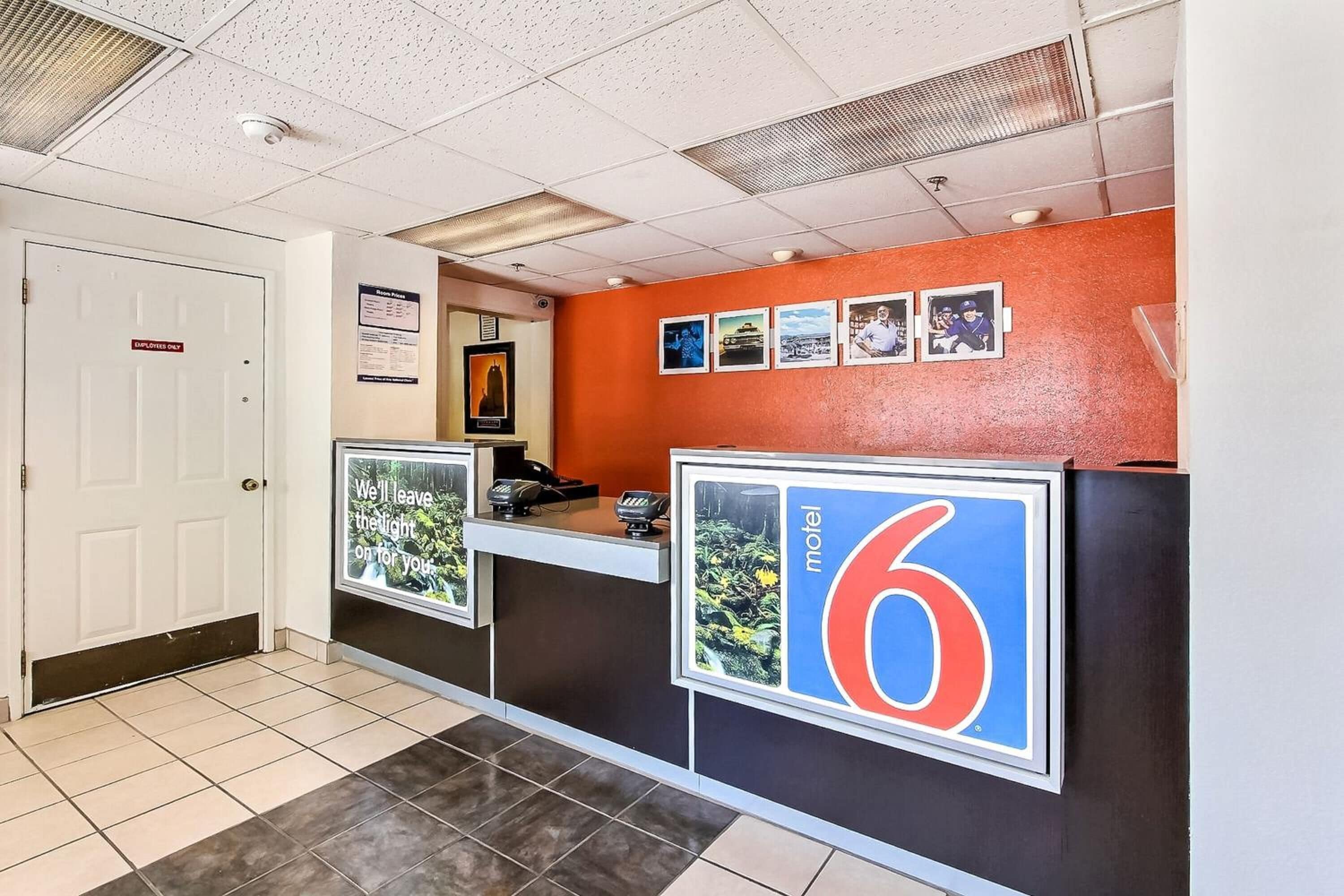 Zona de recepción de un hotel con un letrero con el número '6', un mostrador de recepción, una puerta que indica 'Pago a empleados' y un tablón de anuncios con fotografías.