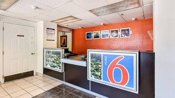 Zona de recepción de un hotel con un letrero con el número '6', un mostrador de recepción, una puerta que indica 'Pago a empleados' y un tablón de anuncios con fotografías.