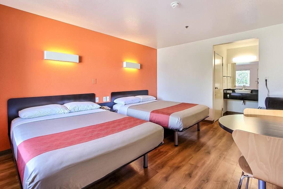 A hotel room with two beds, a wooden floor, a small table, and a bathroom visible through an open door.