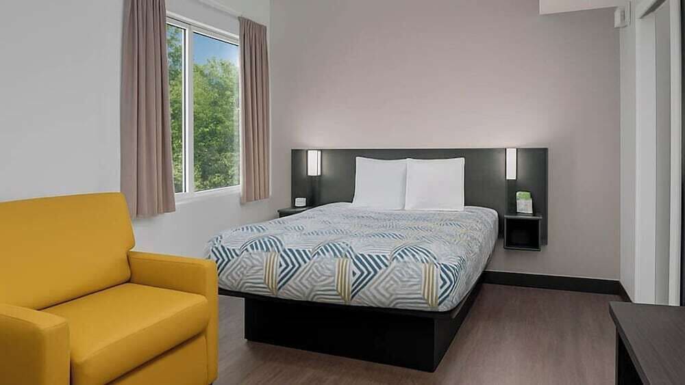 Une chambre d’hôtel avec un lit, un fauteuil jaune, une table de chevet et une fenêtre avec des rideaux.