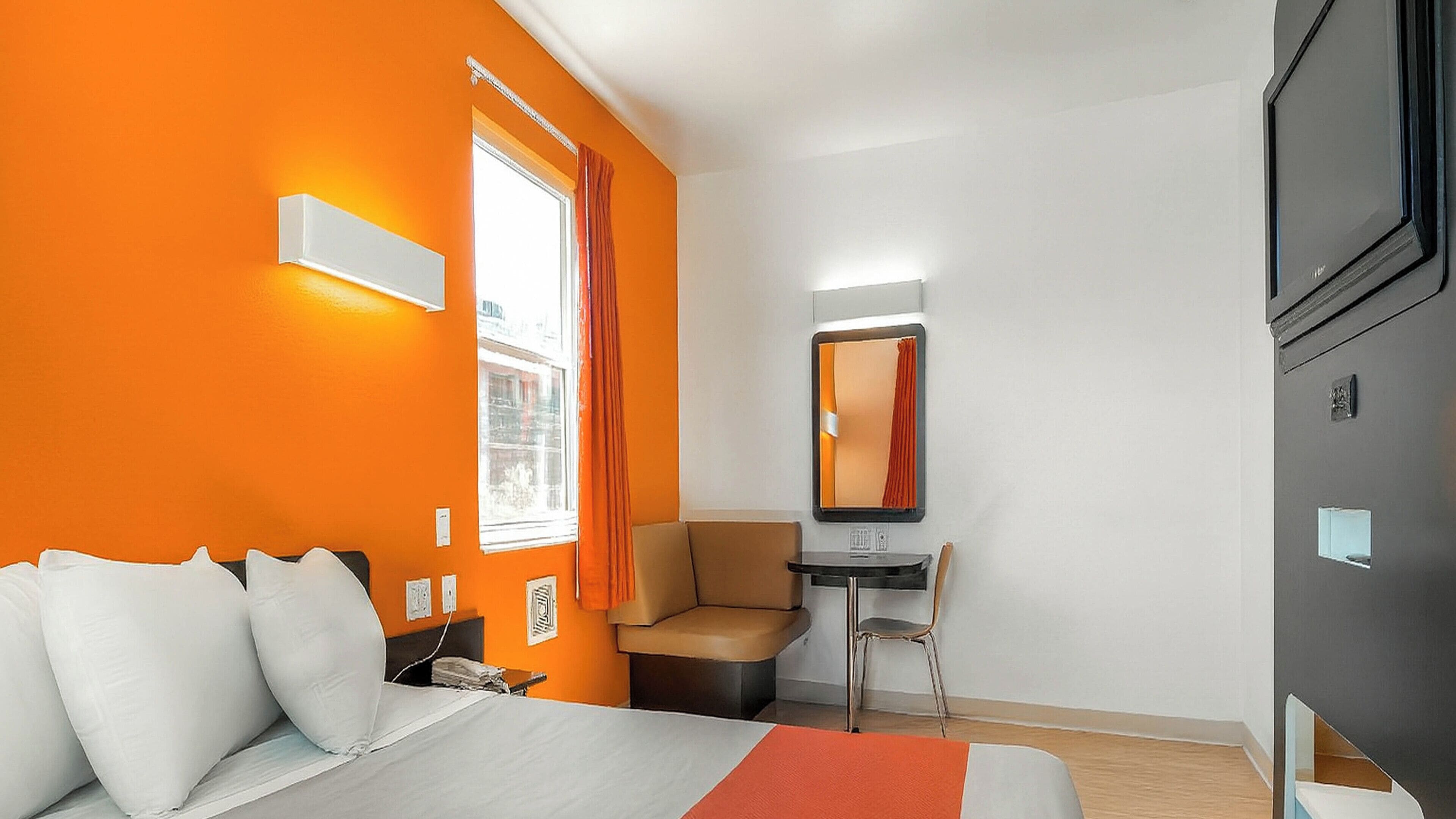 A hotel room with an orange wall, a bed with white bedding, a desk with a chair, and a large mirror.