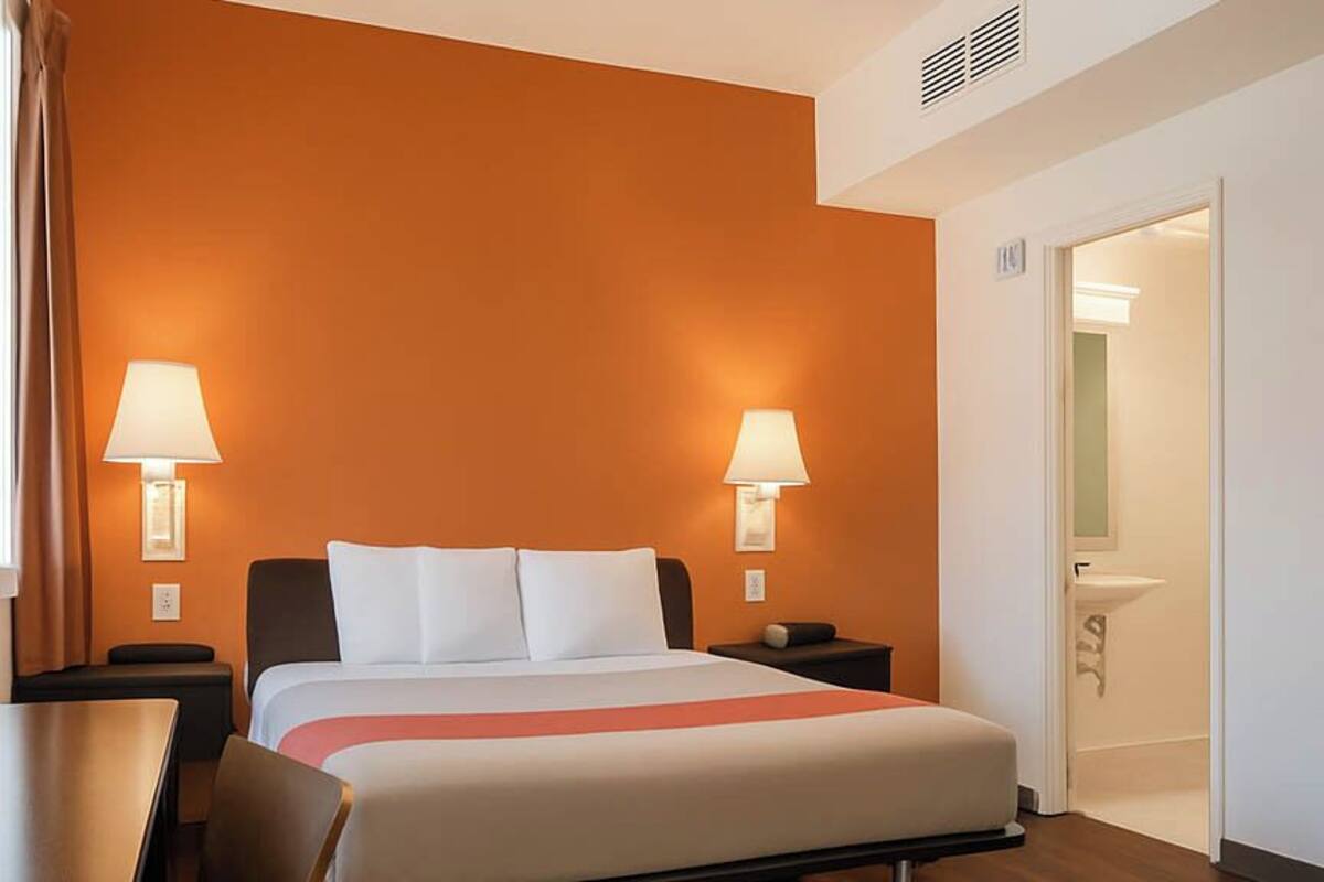 A hotel room with a bed, two bedside lamps, a nightstand, a chair, and a bathroom visible through an open door.
