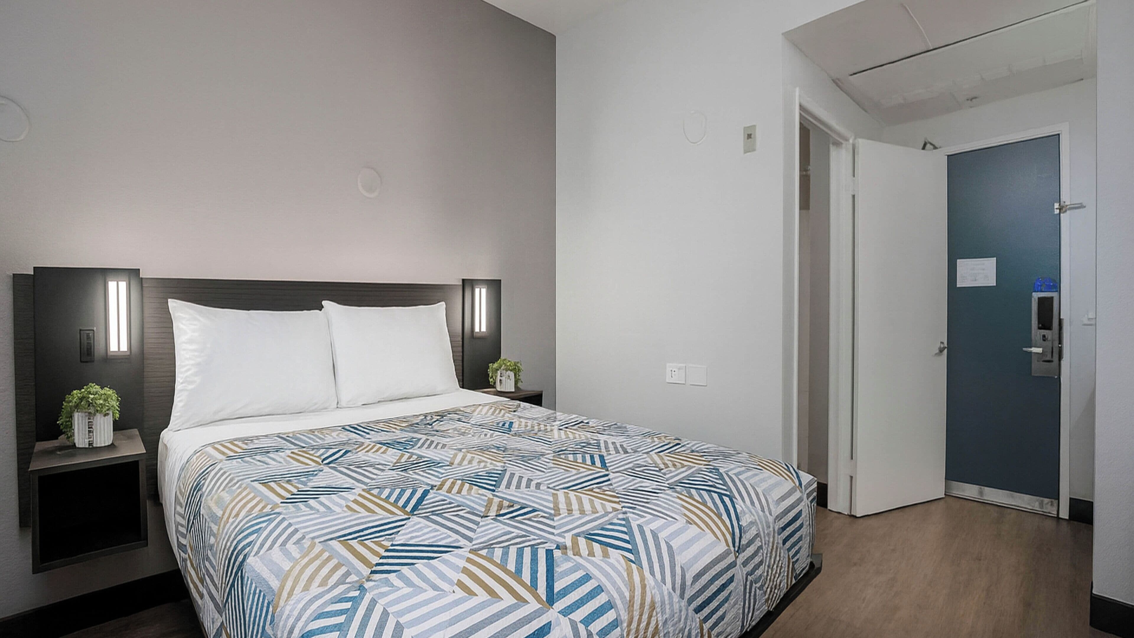 Ein Hotelzimmer mit einem Bett, Nachttischen, einer Wandleuchte, einer Tür und einer kleinen Pflanze.