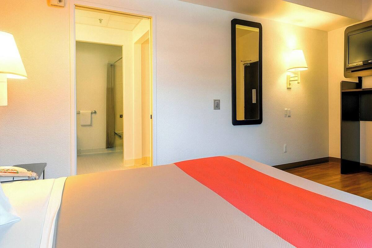 A hotel room with a bed, a TV, a mirror, and a door leading to a bathroom.