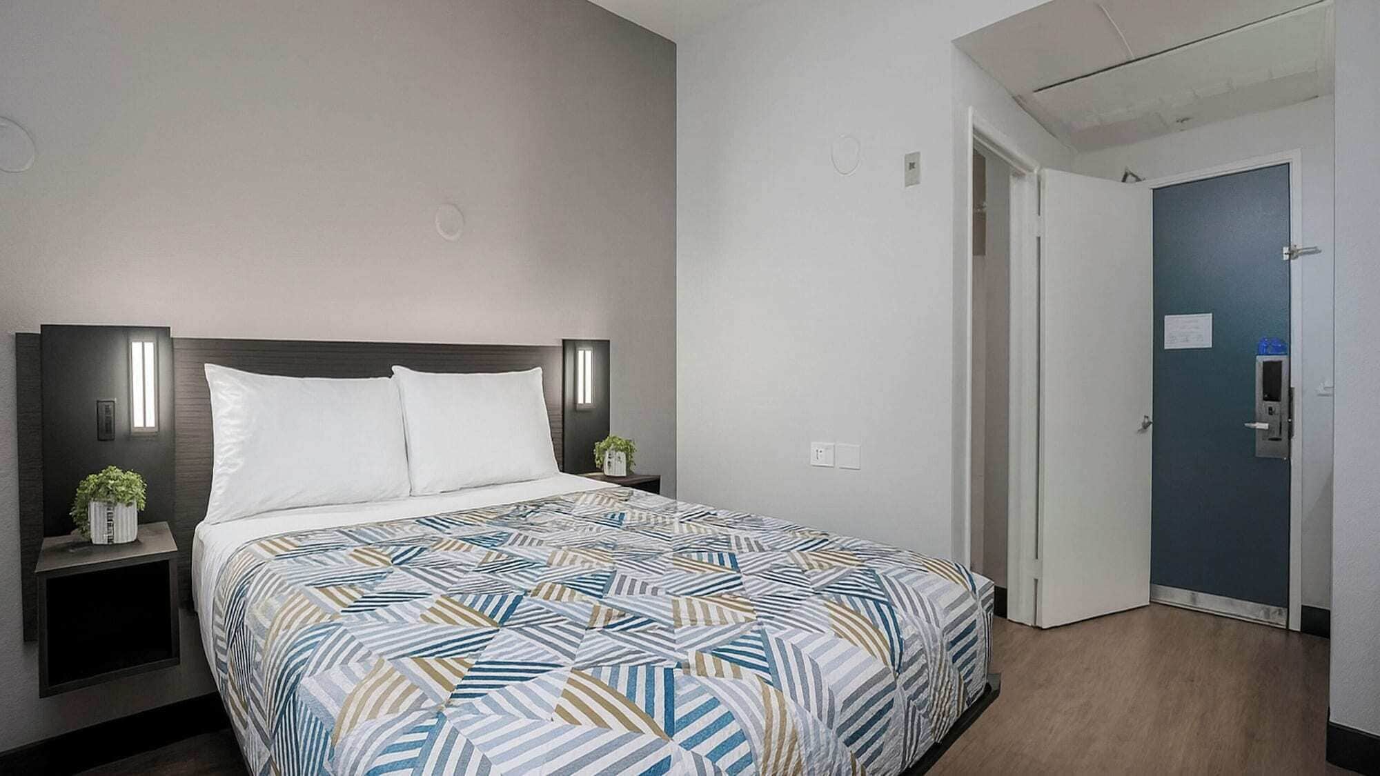 Habitación de hotel con una cama, mesitas de noche, una lámpara de pared y una puerta.