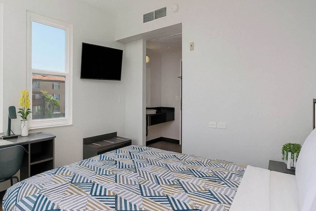 A modern hotel room with a bed, a desk, a chair, a TV, and a window with a view of buildings.