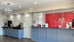 Et hotellets reception område med et moderne design, der inkluderer en disk med rød baggrund, en kaffemaskine og en glasklædt dør, der fører til lobbyen.