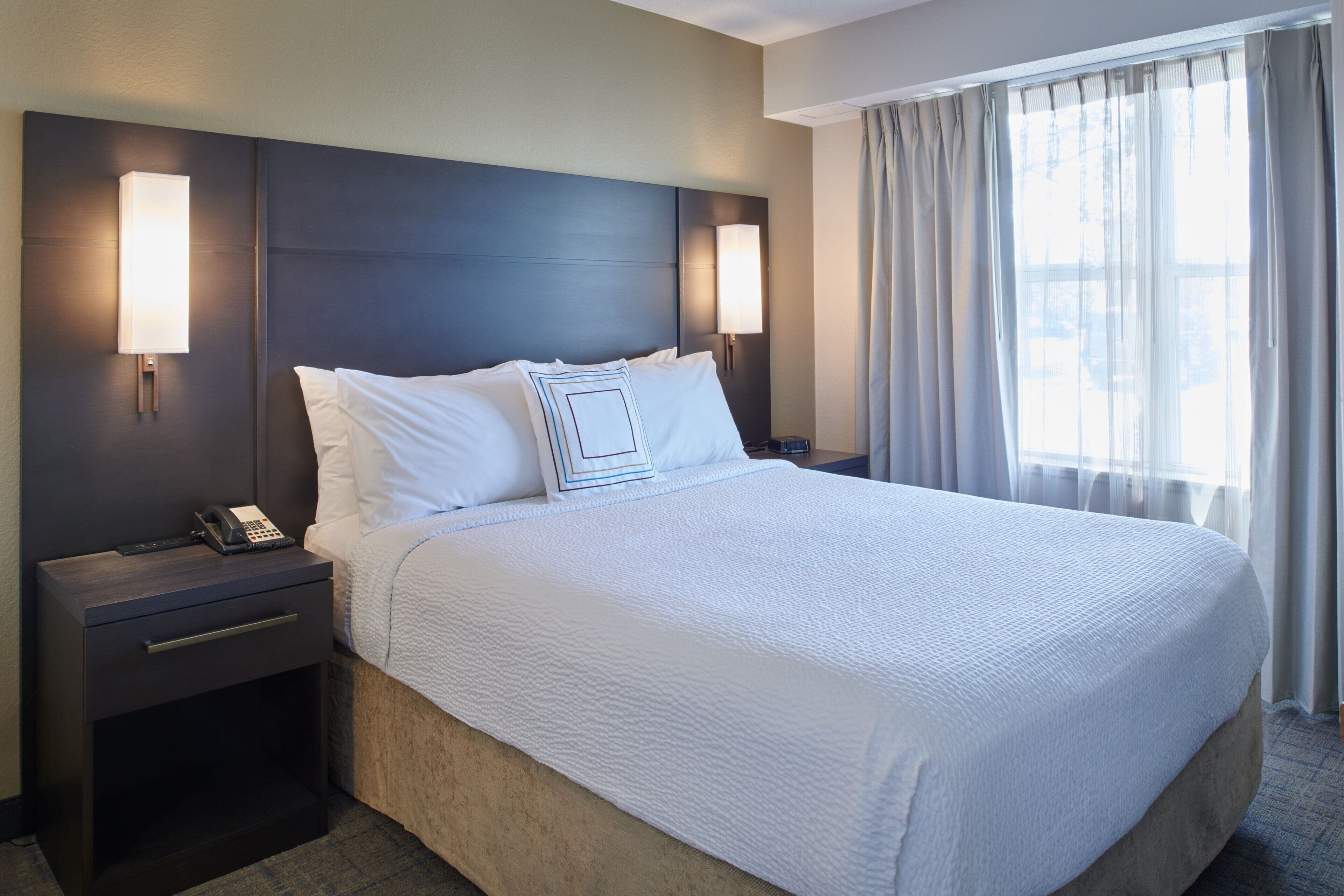 A hotel room with a large bed, two bedside tables with lamps, a bedside phone, and a window with curtains.