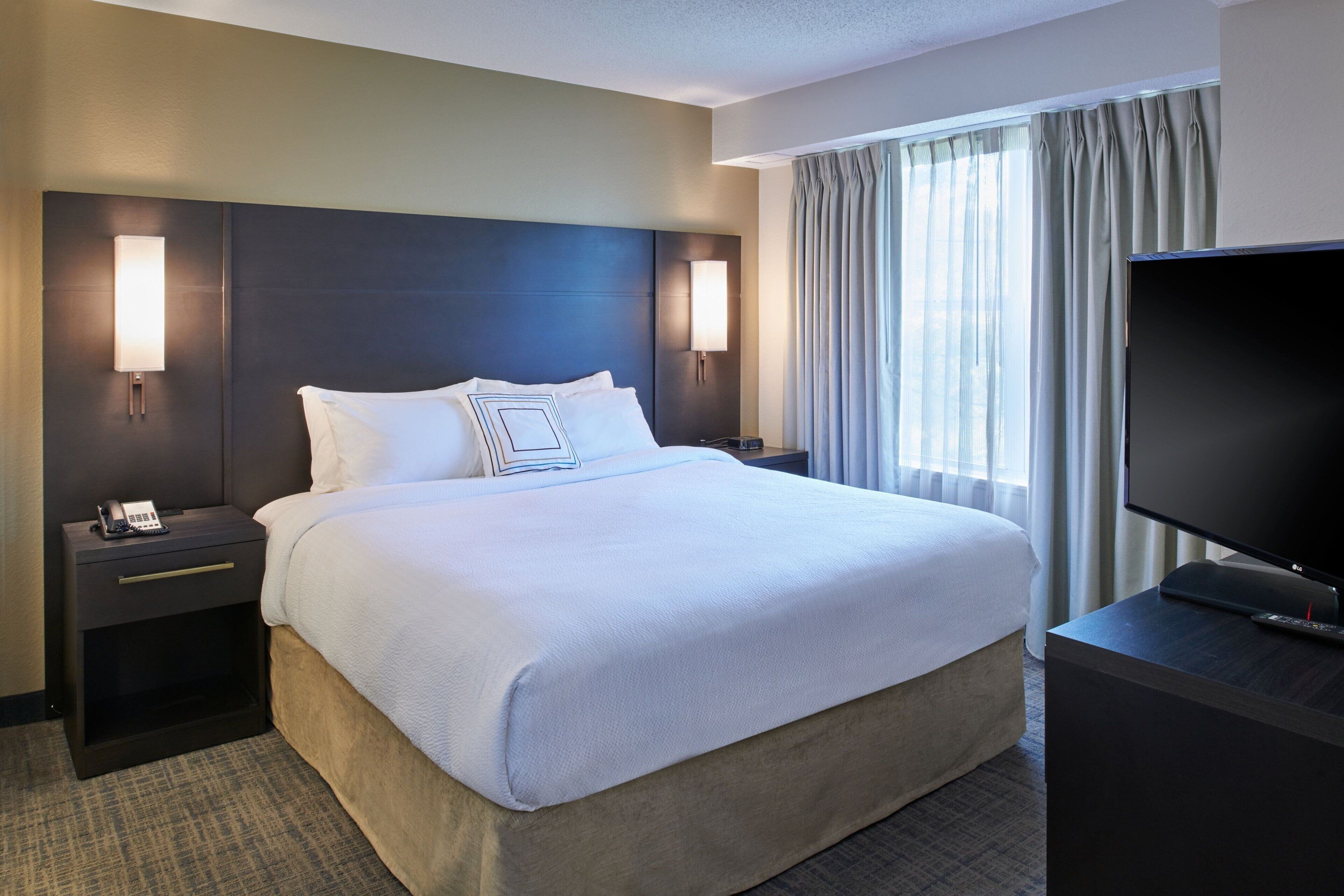A hotel room with a large bed, two bedside tables, a TV, and a window with curtains.
