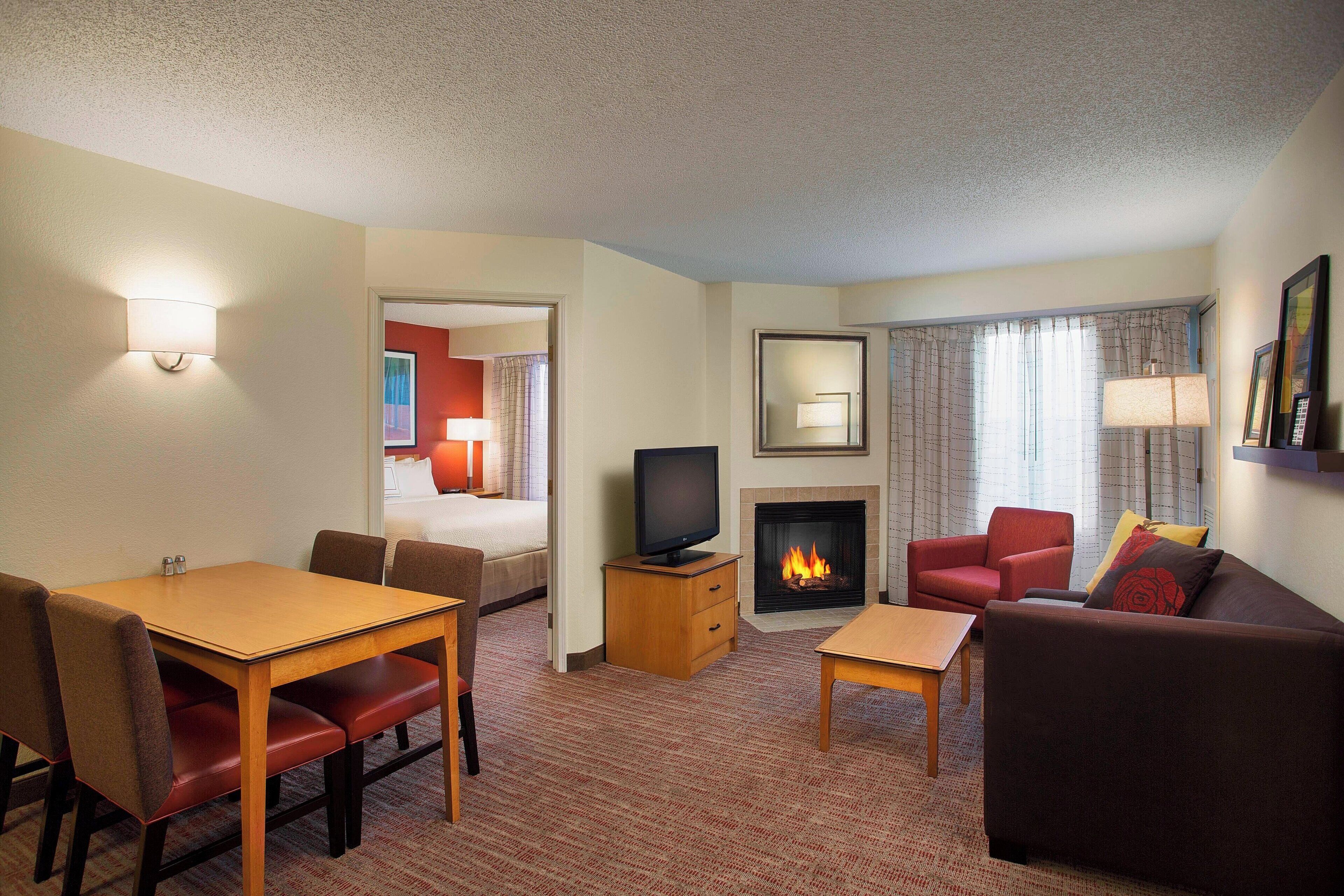 A hotel room with a dining table, chairs, a sofa, a television, a fireplace, and a bed.