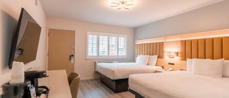 A hotel room with two beds, a desk with a coffee maker, a TV, and a window with blinds.