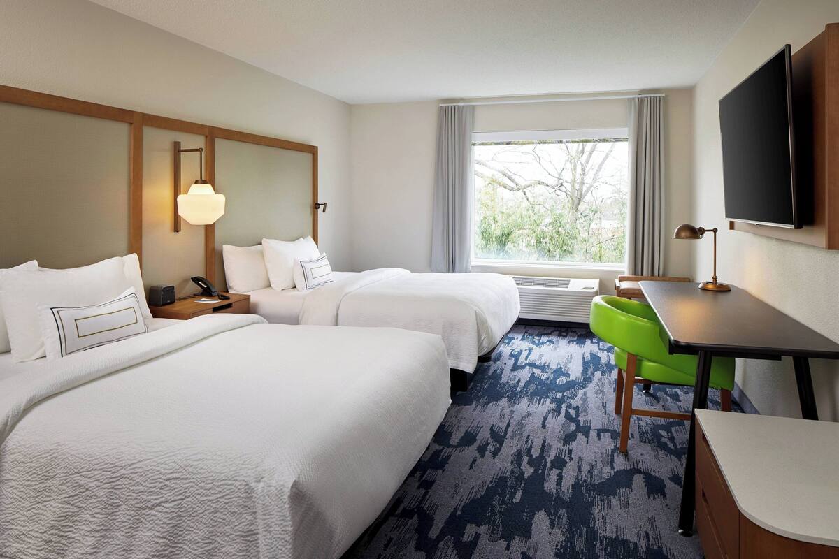 A hotel room with two beds, a desk, a chair, a TV, and a window with a view of trees.