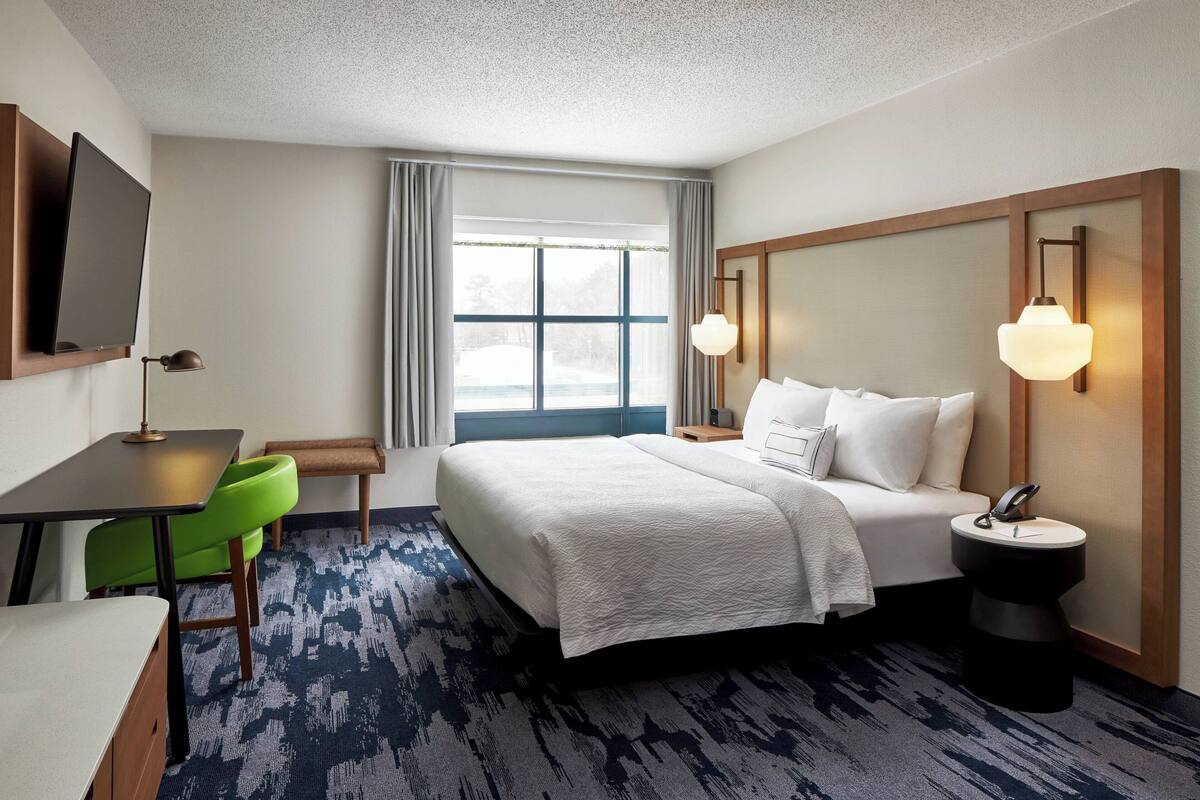 A hotel room with a bed, a desk, a chair, a nightstand, a TV, and a window with curtains.