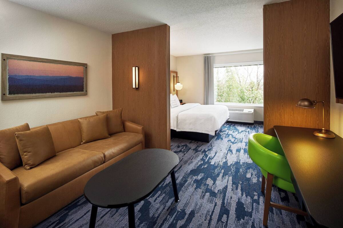 A hotel room with a brown leather sofa, a small table, a green chair, a bed with white linens, a large window, and a painting on the wall.