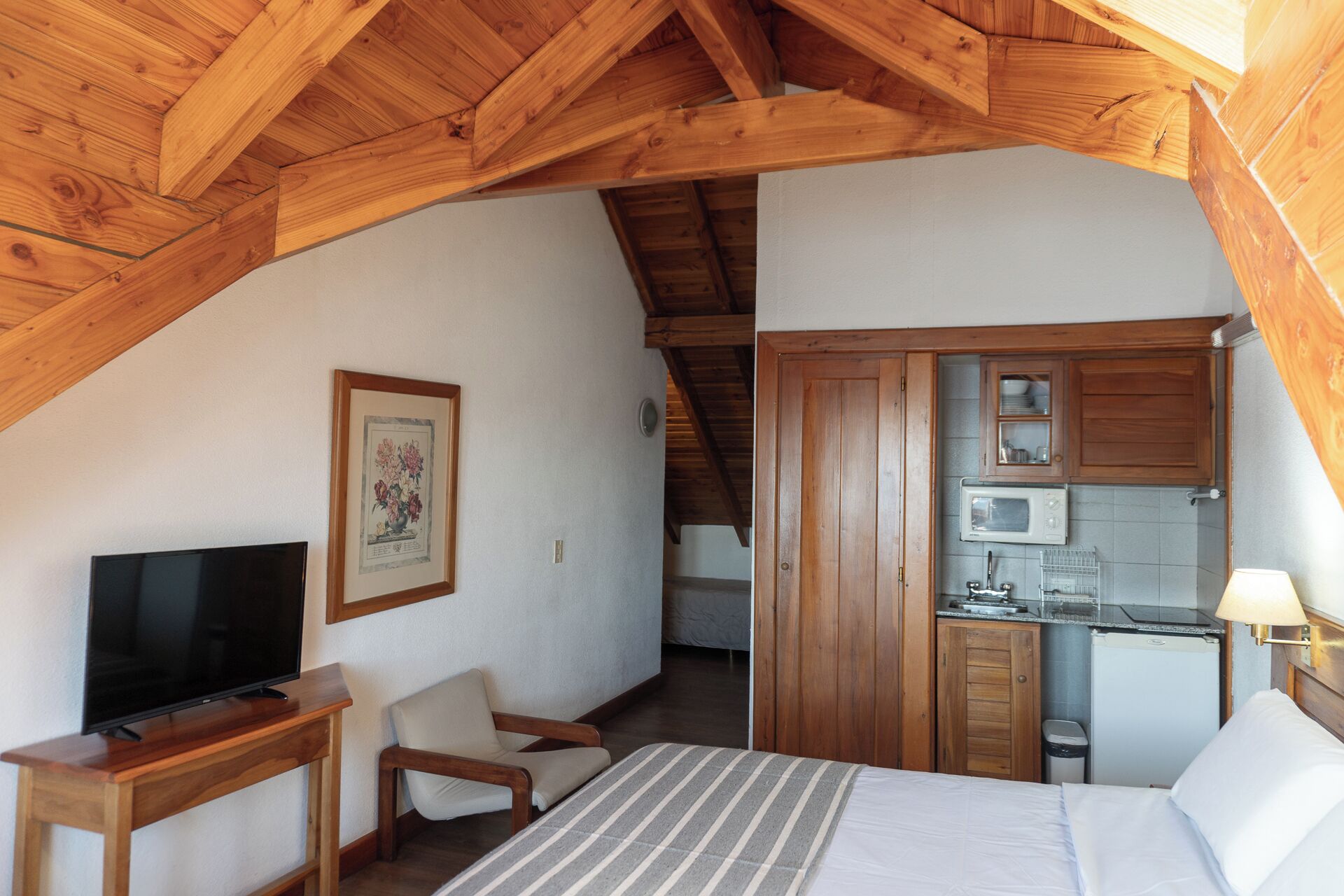 Ein gemütliches Schlafzimmer im Dachgeschoss mit Holzbalkendecke, Fernseher, Bett, Stuhl und einer Küchenzeile.