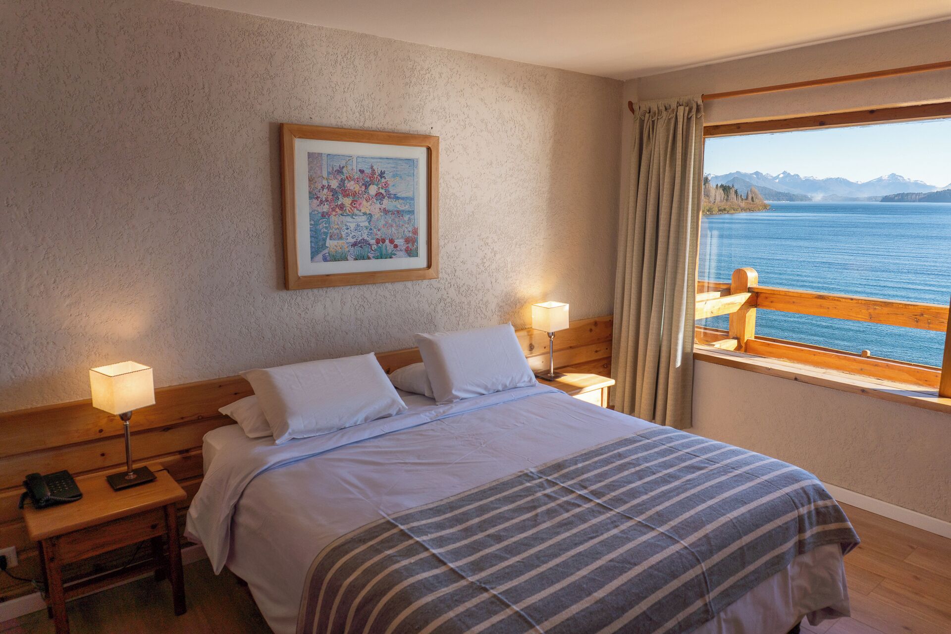 A hotel room with a bed, bedside lamps, a painting, and a window with a view of the sea and mountains.