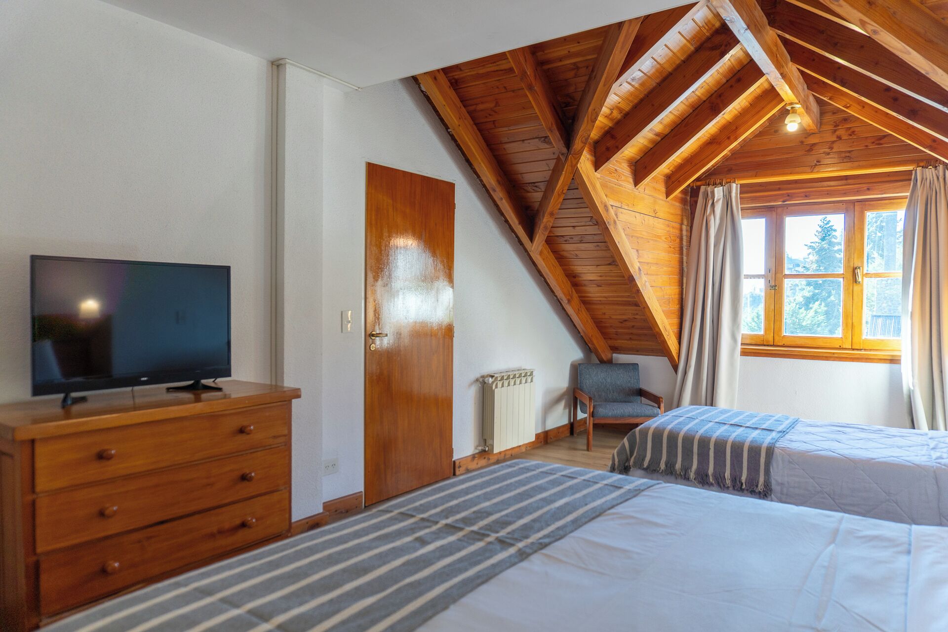 Een slaapkamer met schuine wand, een televisie op een houten dressoir, een stoel en een raam met gordijnen.