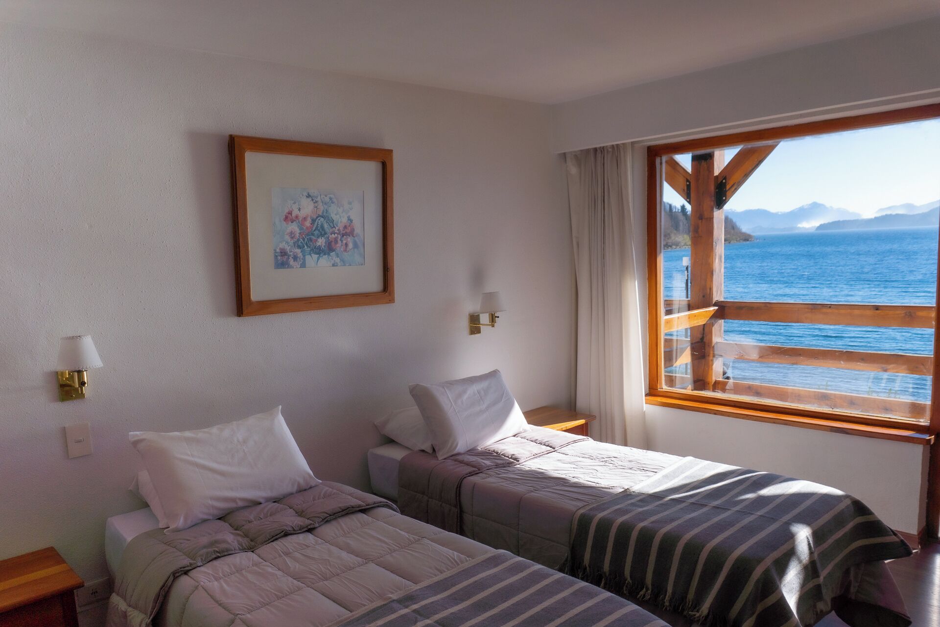 A room with two beds, a framed picture on the wall, a window with a view of the sea and mountains, and a small table with a lamp.