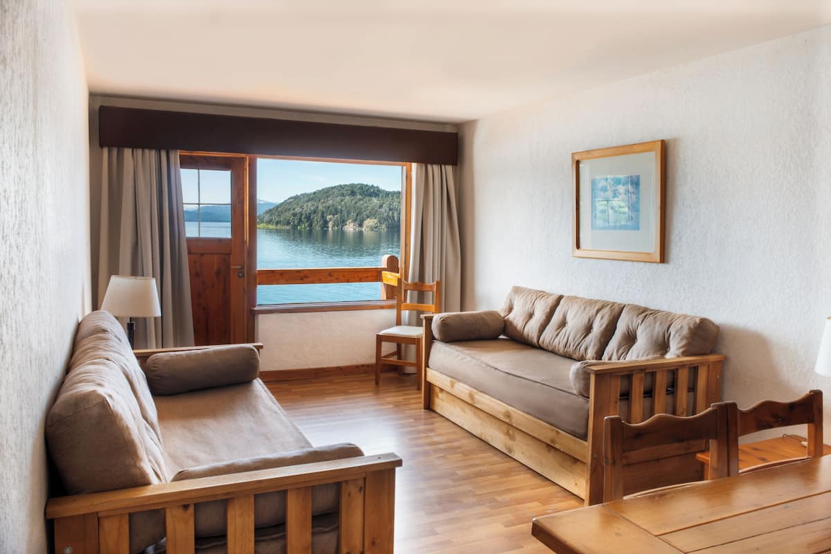 Une pièce chaleureuse avec un plancher en bois, un canapé, un fauteuil et une table. Une fenêtre offre une vue sur un lac et des collines.