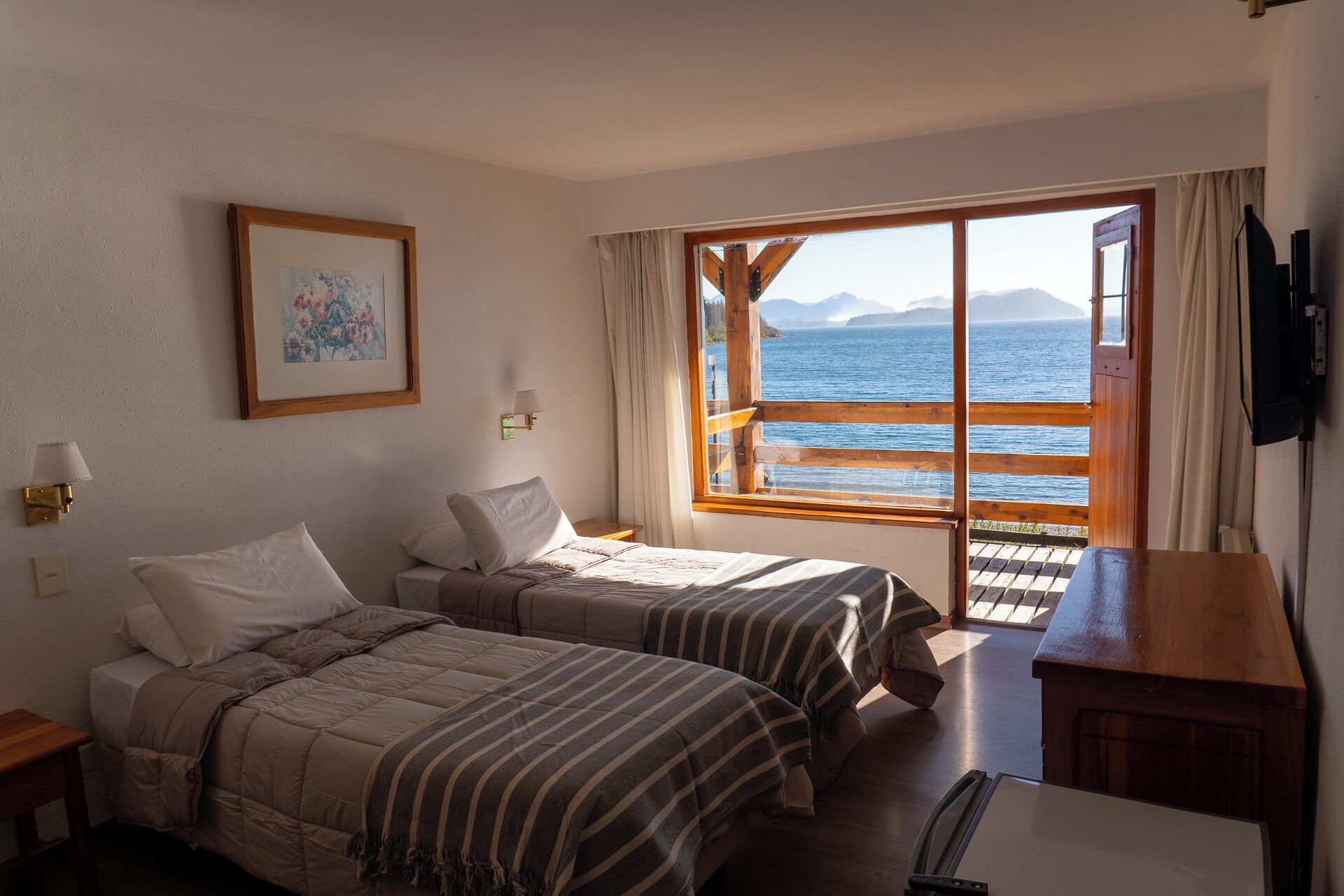 Une chambre d’hôtel avec deux lits, un bureau en bois, une télévision et une grande fenêtre donnant sur la mer et les montagnes.