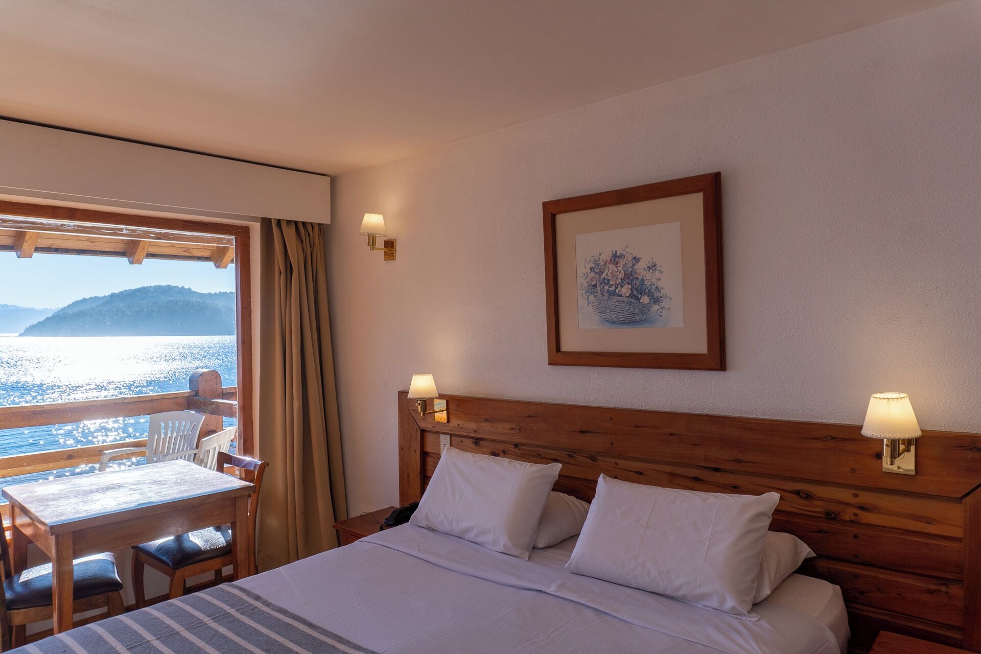 A hotel room with a bed, two bedside lamps, a framed picture on the wall, a wooden headboard, a table with chairs, and a view of the sea through a window.